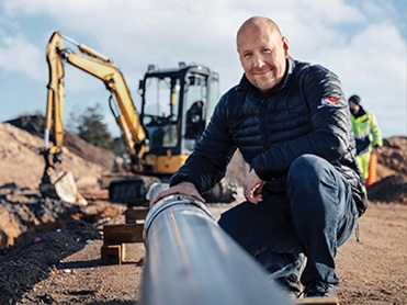 Showing a man in focus on a building site at a large pipe smiling into the camera with a blurred background of a working man and a digging machine