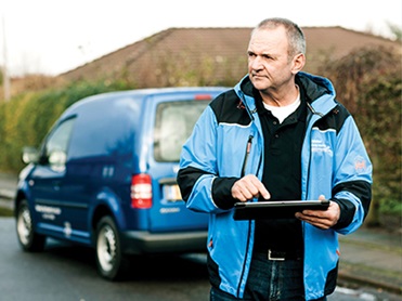 A man in blue jacket outside with his car in the background holding a tablet, reading water consumption remotely 